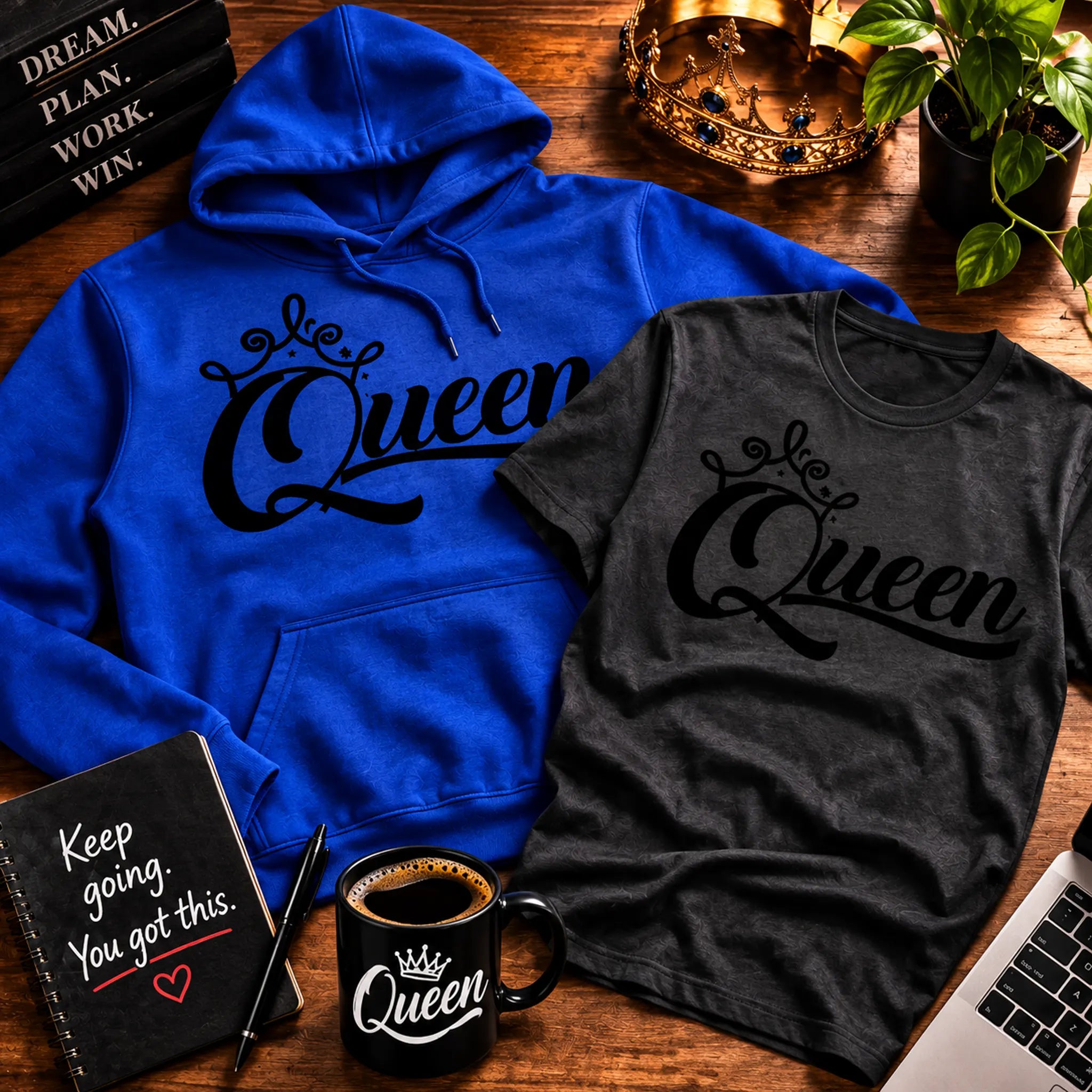 A blue hoodie and a dark gray t-shirt, both featuring a black 'Queen' graphic with a crown, arranged in a styled flat-lay on a wooden desk with a coffee mug, notebook, laptop, and plant.