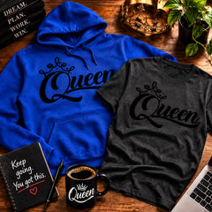 A blue hoodie and a dark gray t-shirt, both featuring a black 'Queen' graphic with a crown, arranged in a styled flat-lay on a wooden desk with a coffee mug, notebook, laptop, and plant.