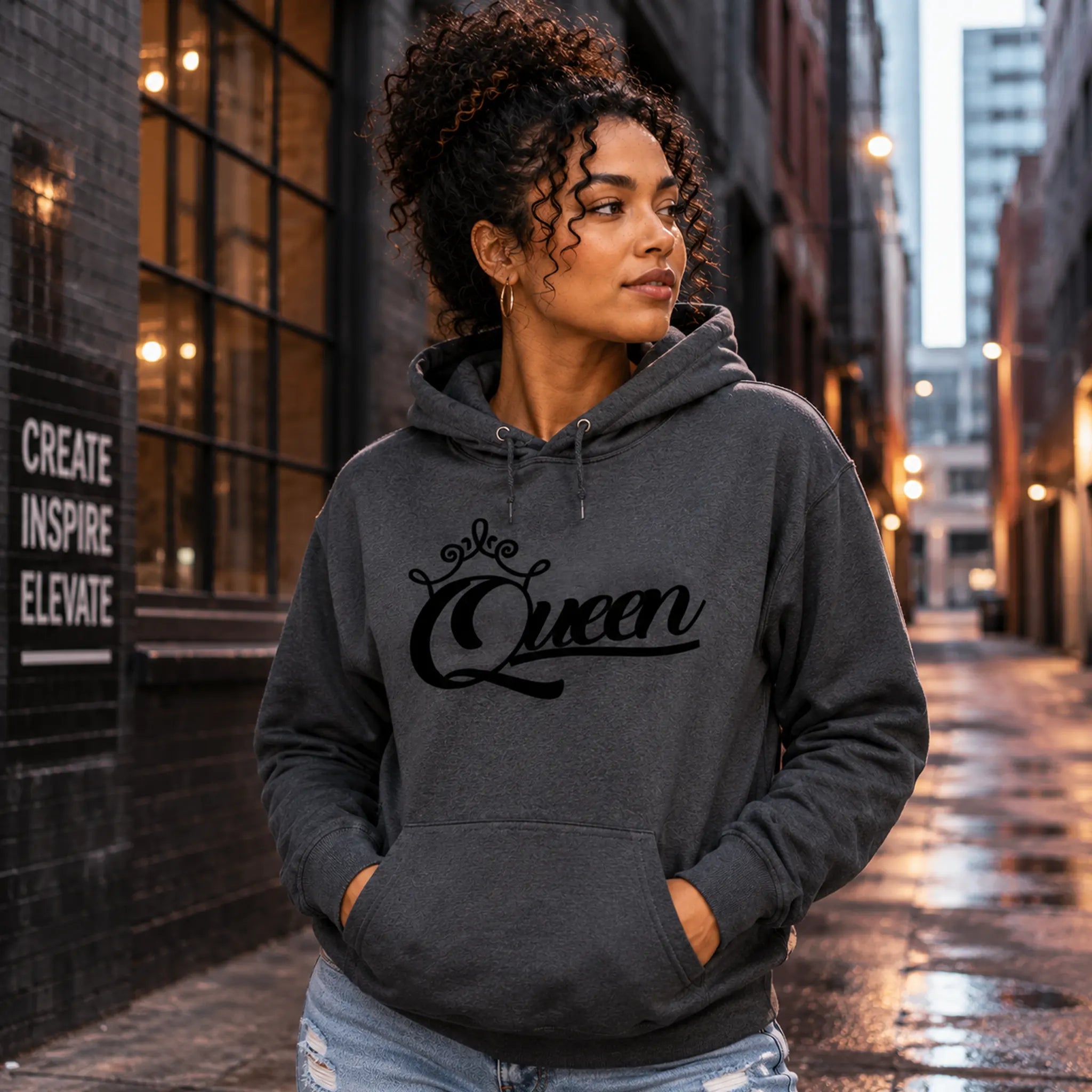 Woman wearing Queen Dark Heather pullover hoodie with black crown script design in an urban alley setting – Black Heart Tees motivational apparel.