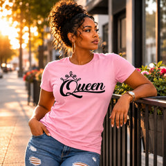 A woman wearing a light pink crewneck t-shirt with 'Queen' written in black script and a small crown graphic, paired with distressed blue jeans, leaning against a black metal railing on a city sidewalk during golden hour.