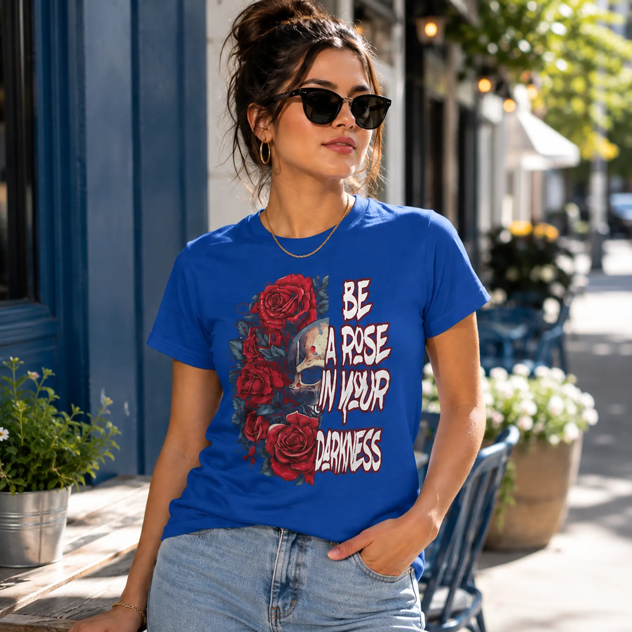 Woman wearing Be A Rose In Your Darkness True Royal graphic tee with skull and red roses design in an outdoor café setting – Black Heart Tees motivational apparel