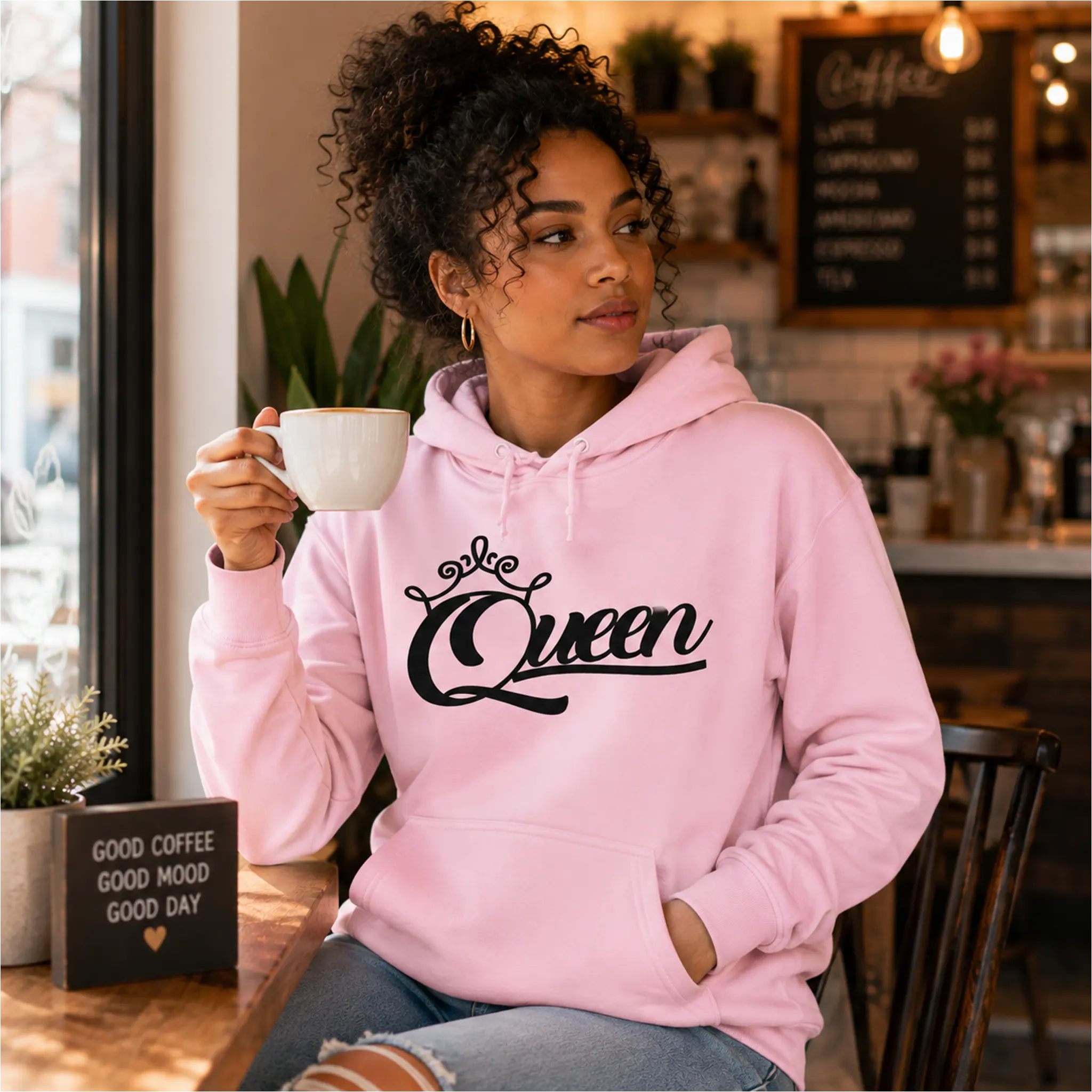 Woman wearing Queen light pink pullover hoodie with black crown script design in a cozy coffee shop – Black Heart Tees motivational apparel.