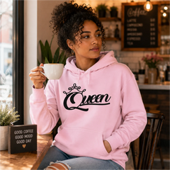 Woman wearing Queen light pink pullover hoodie with black crown script design in a cozy coffee shop – Black Heart Tees motivational apparel.