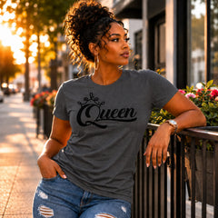 A woman wearing a dark gray graphic t-shirt with 'Queen' written in black script under a crown, paired with distressed blue jeans, leaning on a black metal railing in an outdoor urban setting during golden hour.