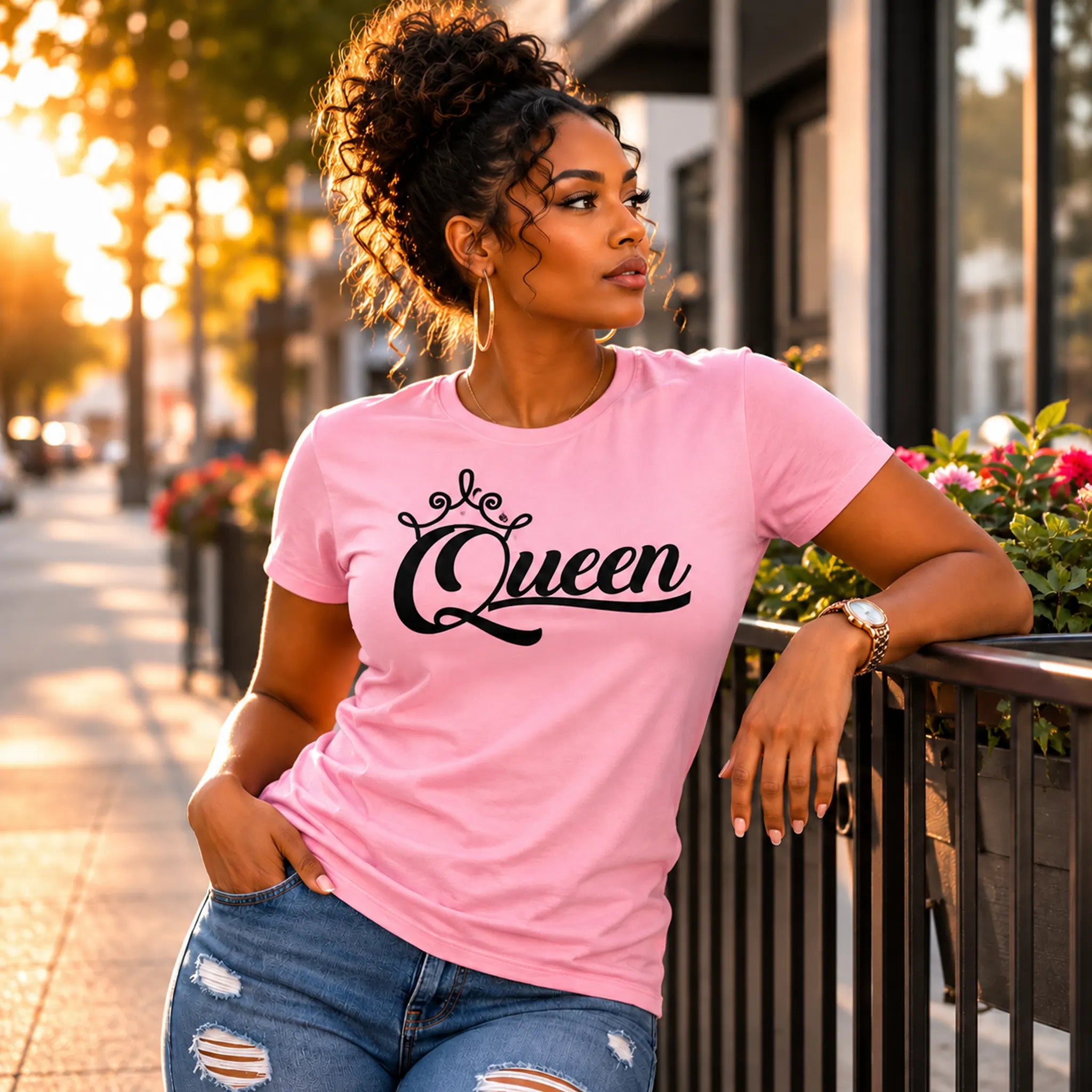 A woman wearing a light pink crewneck t-shirt with 'Queen' written in black script and a small crown graphic, paired with distressed blue jeans, leaning against a black metal railing on a city sidewalk during golden hour.