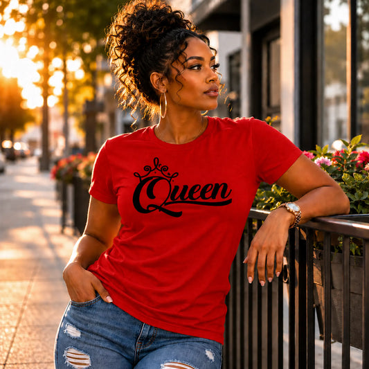 A woman wearing a bright red crewneck t-shirt with 'Queen' written in black script across the chest, posing outdoors on a city sidewalk at sunset.