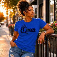 A woman wearing a bright blue crewneck t-shirt with 'Queen' written in black script across the chest, posing outdoors on a city sidewalk at sunset.