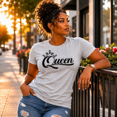 A woman wearing a light gray crewneck t-shirt with 'Queen' written in black script and a small crown graphic, paired with distressed blue jeans, posing outdoors on a city sidewalk during golden hour.