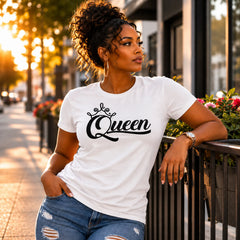 A woman wearing a white crewneck t-shirt with 'Queen' written in black script and a small crown graphic, paired with distressed blue jeans, posing outdoors on a city sidewalk during golden hour.